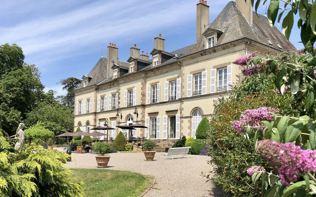 Terrasse du Château d’Ygrande près de Vichy, bordée de massifs fleuris, avec tables sous parasols devant la façade aux volets blancs.