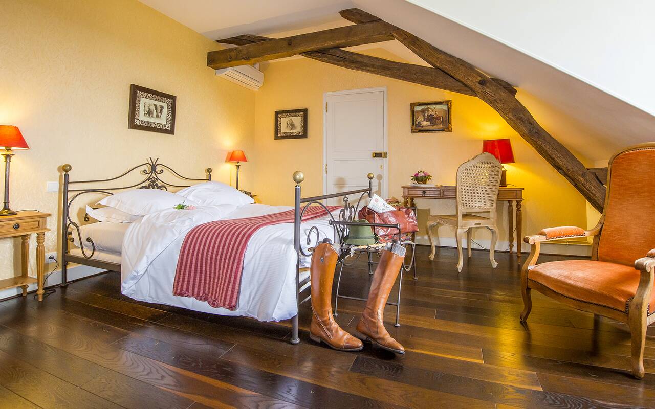 Chambre du Château d’Ygrande près de la forêt de Tronçais : lit en fer forgé sous les poutres apparentes, bottes d’équitation et chapeau au pied du lit.