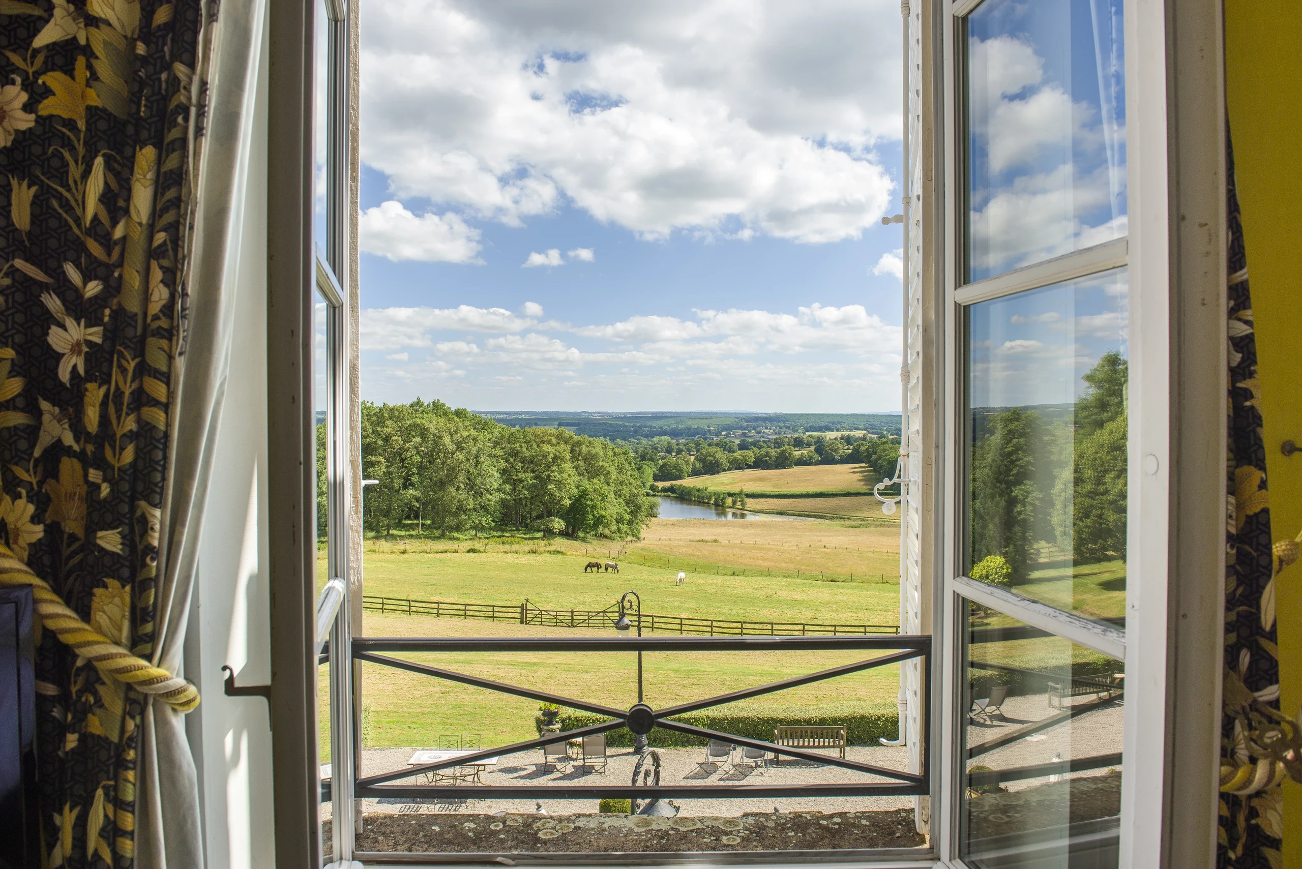 Vue sur les prairies, les chevaux et un étang depuis la fenêtre ouverte d’une chambre d’hôtes en Auvergne.
