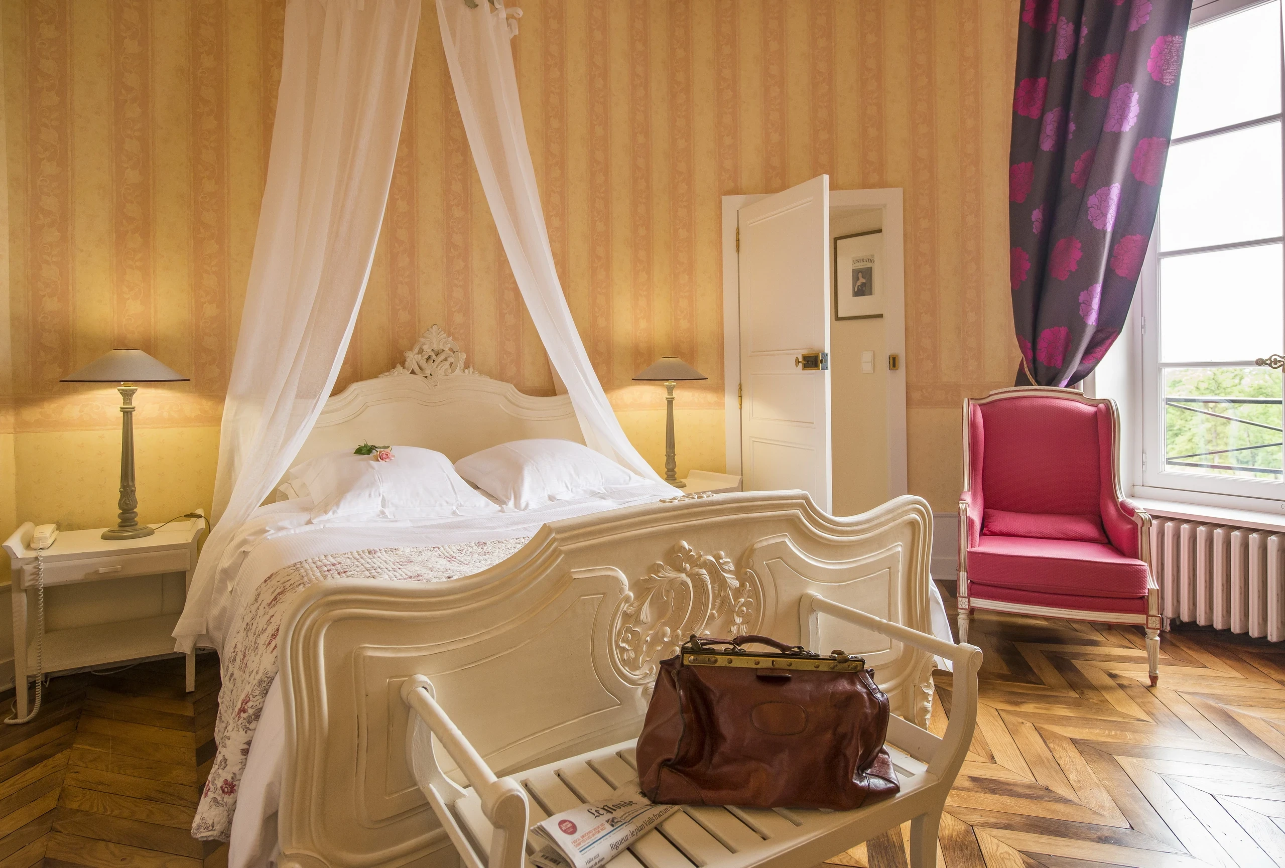 Lit sculpté surmonté de voilages et fauteuil rose près de la fenêtre dans une chambre d’hôtes du Château d’Ygrande en Auvergne.