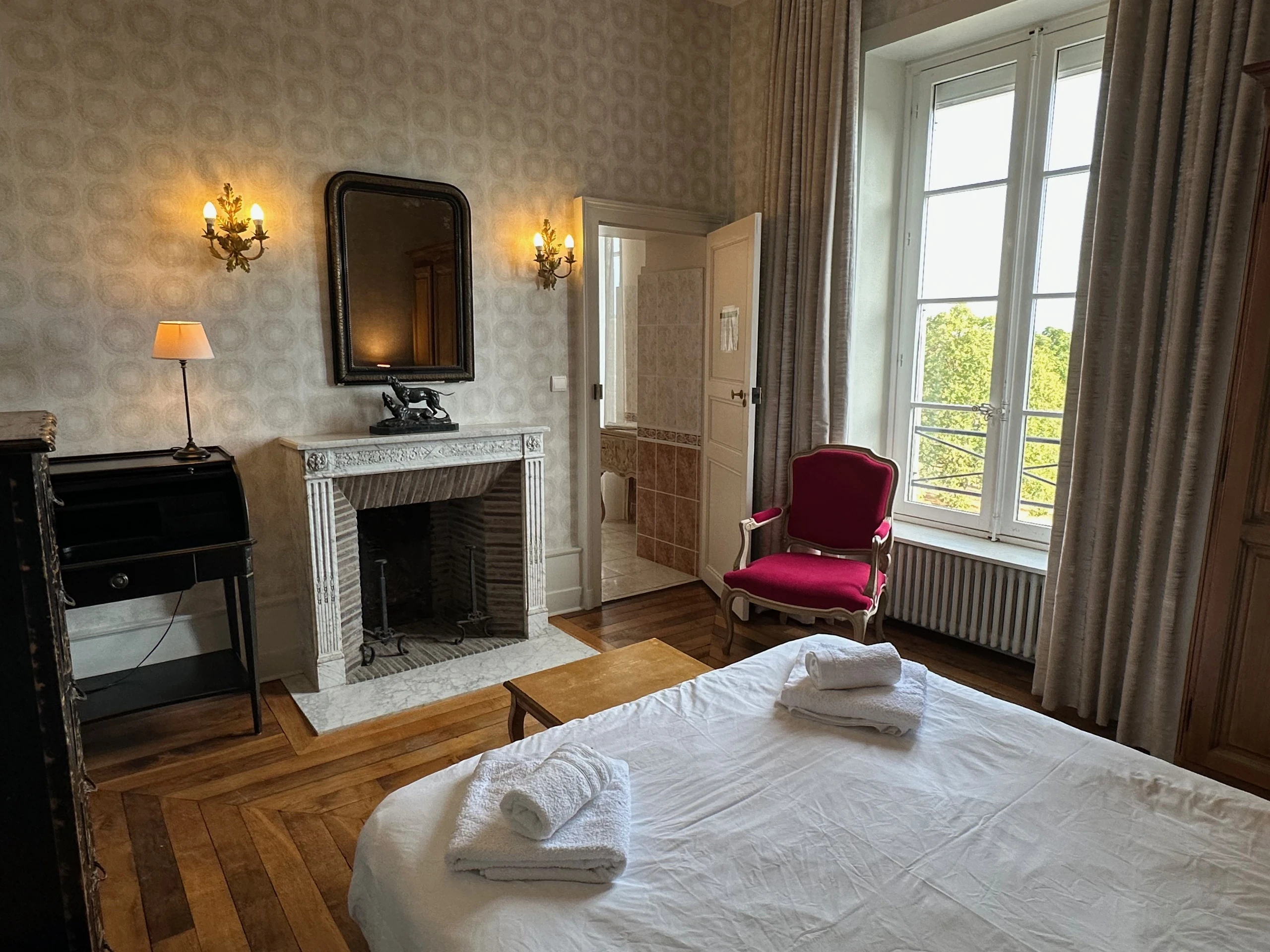 Cheminée en marbre, miroir et fauteuil rouge près d’une grande fenêtre dans une chambre d’hôtes en Auvergne.
