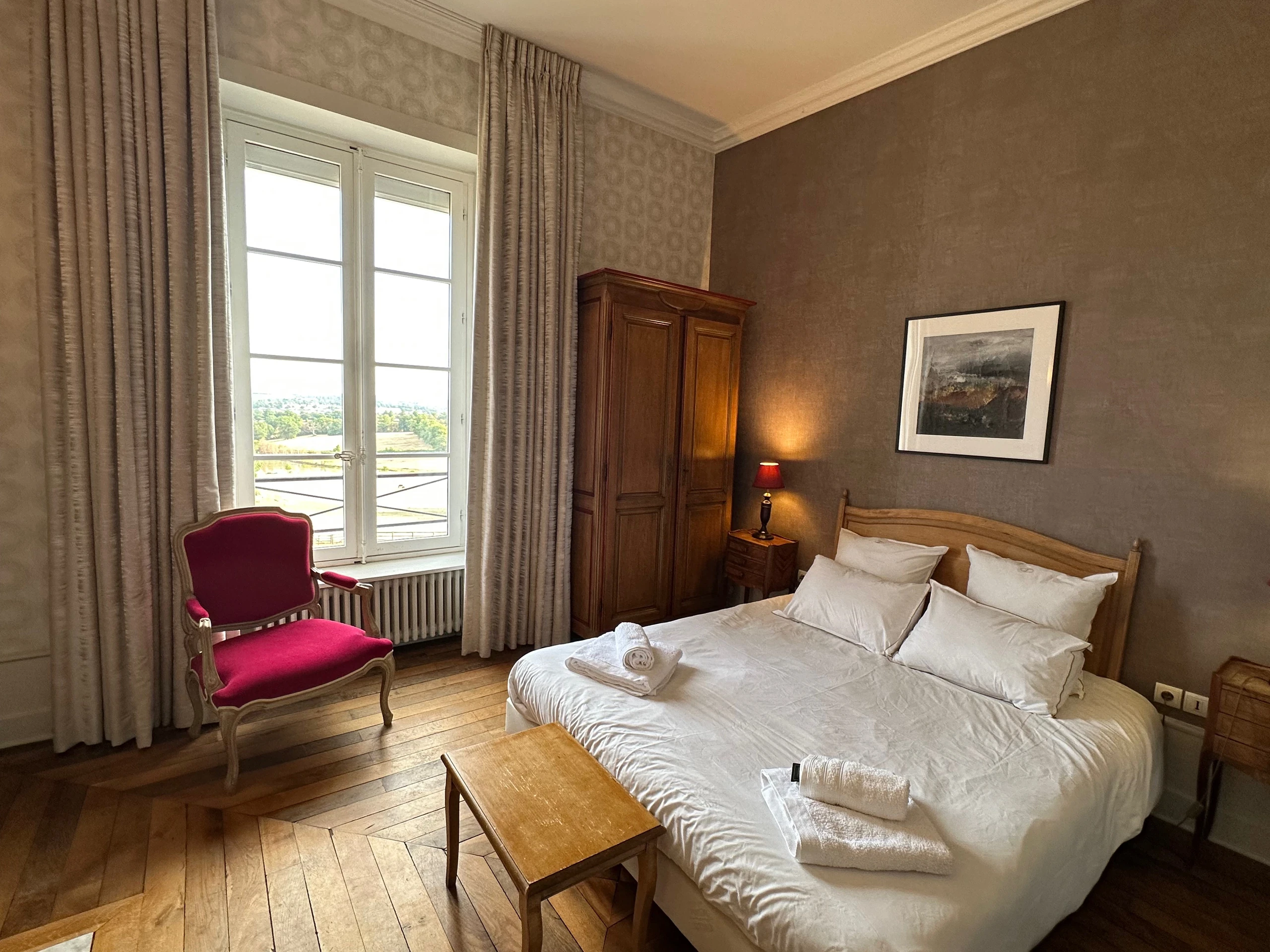 Lit double, armoire en bois et fauteuil rouge près de la fenêtre dans une chambre d’hôtes du Château d’Ygrande en Auvergne.
