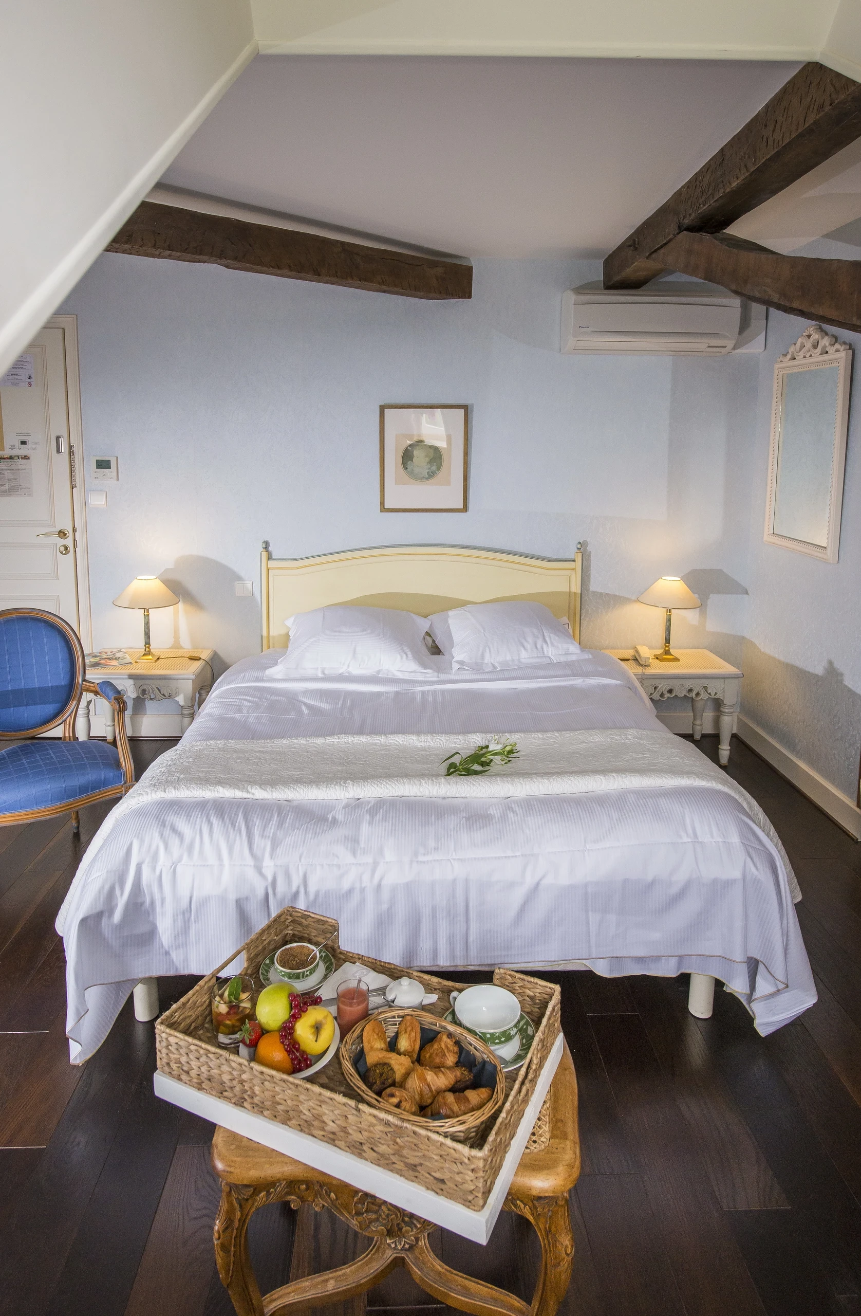 Plateau de petit-déjeuner au premier plan devant un lit double sous des poutres apparentes, dans une chambre d’hôtes en Auvergne.
