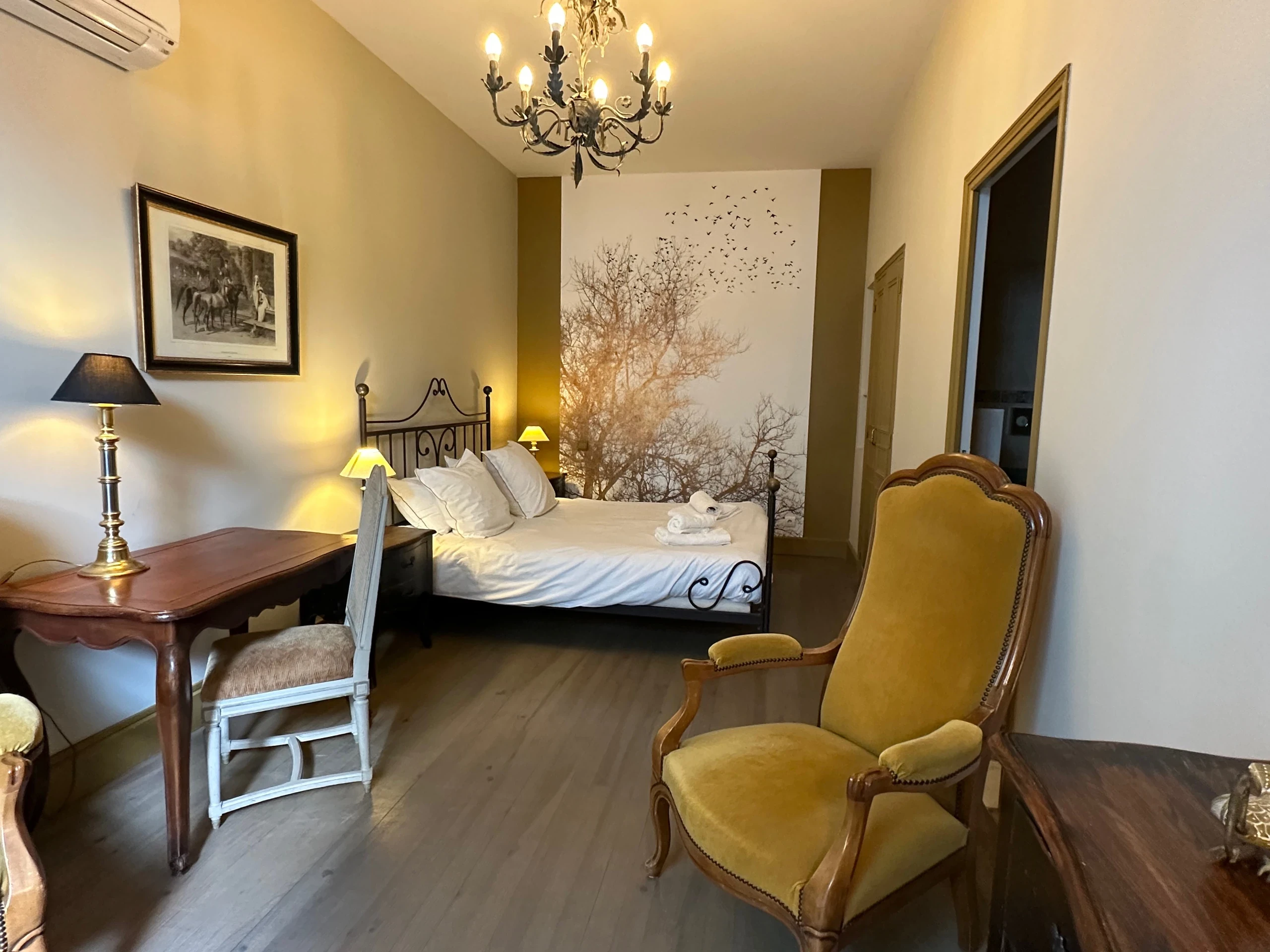 Lit en fer forgé et fauteuil jaune devant un décor mural d’arbre et d’oiseaux dans une chambre d’hôtes en Auvergne.