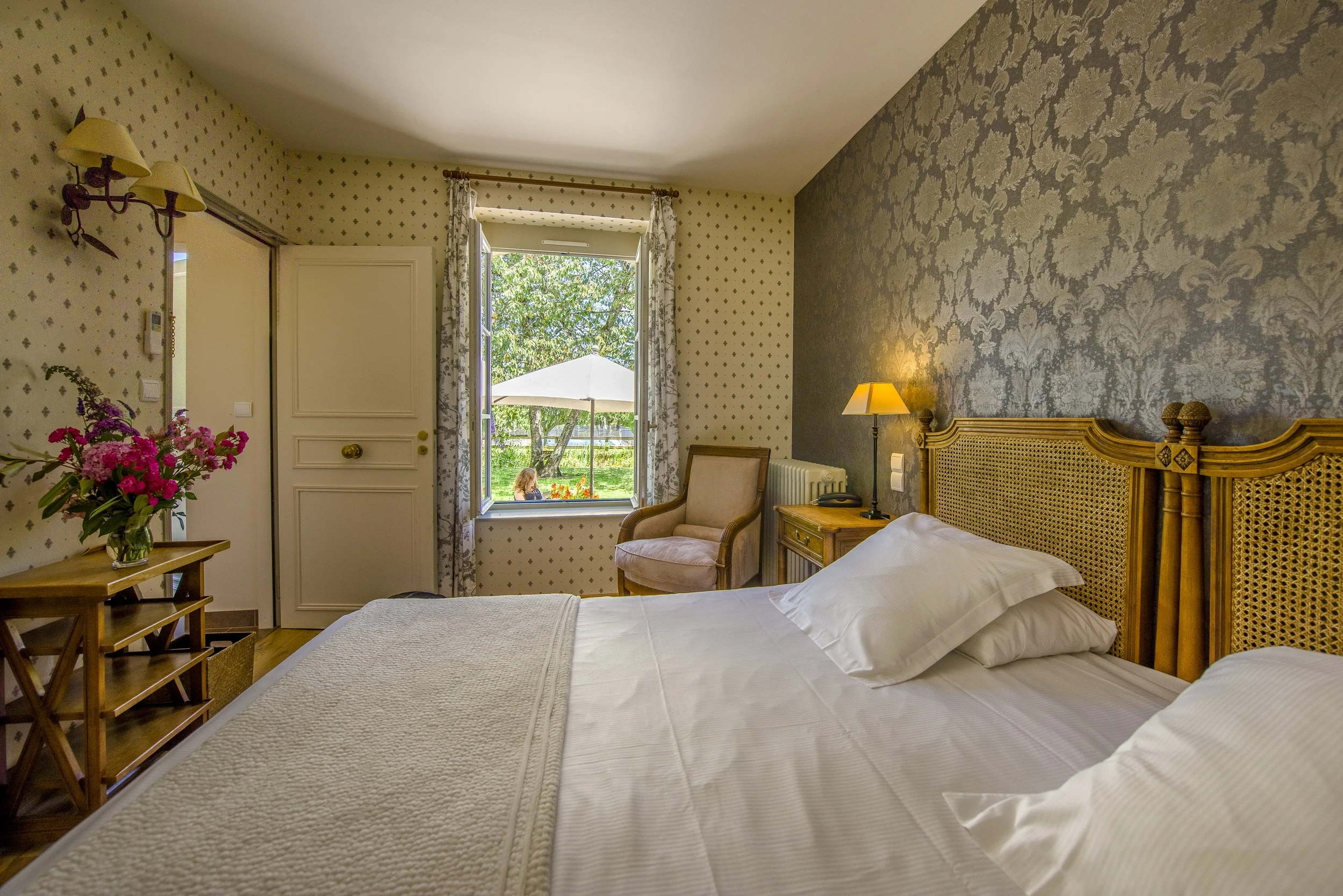 Fenêtre ouverte sur le jardin, fauteuil et tête de lit cannée dans une chambre d’hôtes en Auvergne.