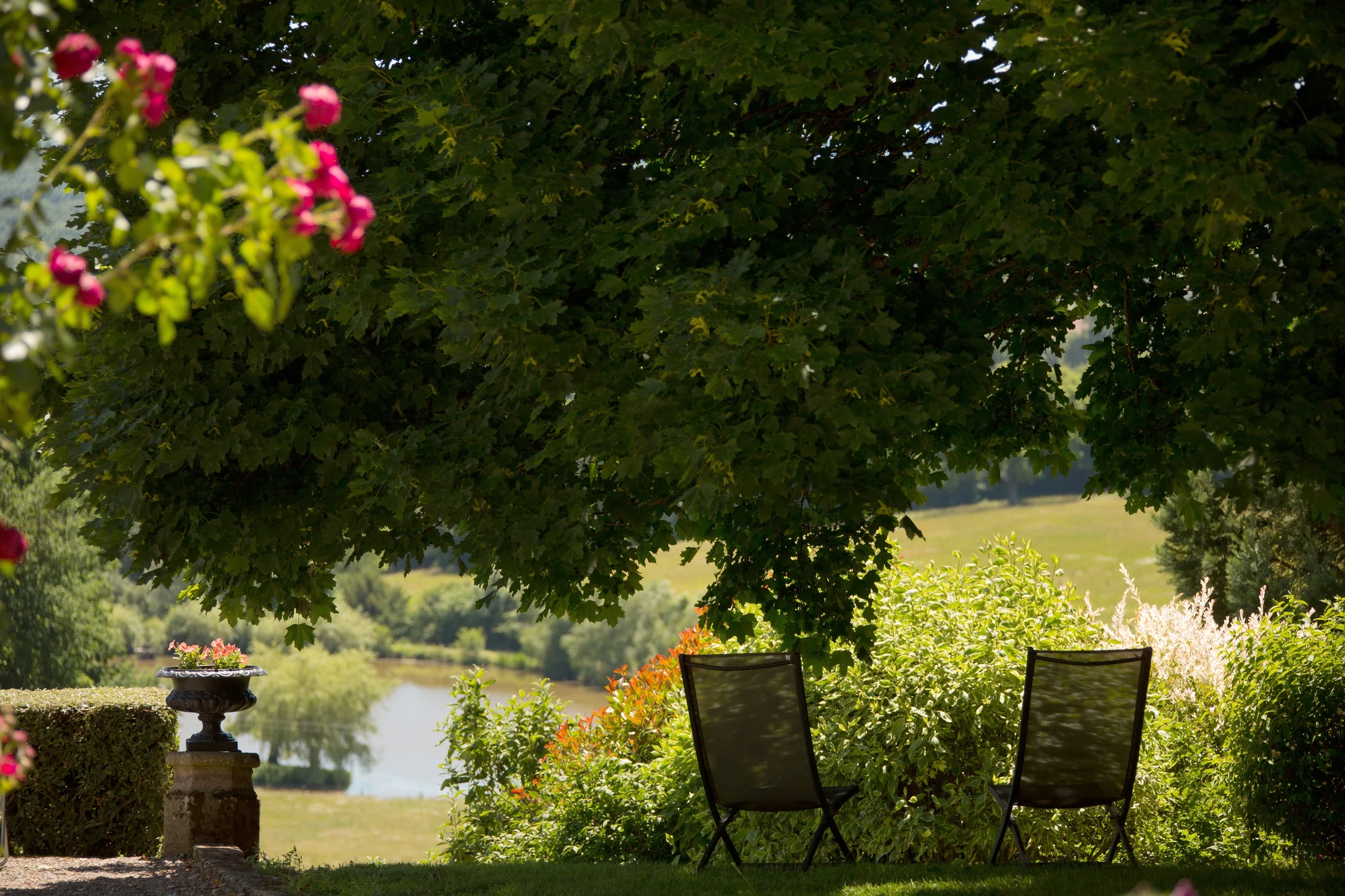 Deux chaises à l’ombre d’un arbre, face à un étang, dans le jardin d’une chambre d’hôtes en Auvergne.