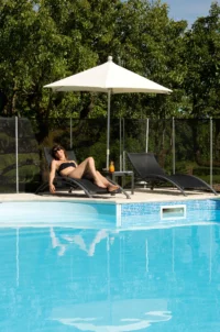 Femme allongée sur un transat sous un parasol au bord de la piscine d’une chambre d’hôtes en Auvergne.